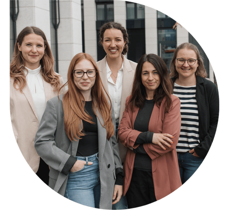 Fünf lächelnde Frauen stehen in der elegante Kleidung vor einem modernen Bürogebäude.