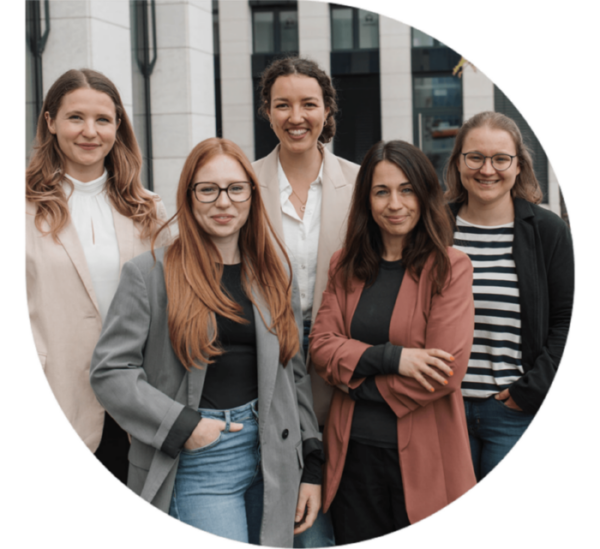 Fünf lächelnde Frauen stehen in der elegante Kleidung vor einem modernen Bürogebäude.