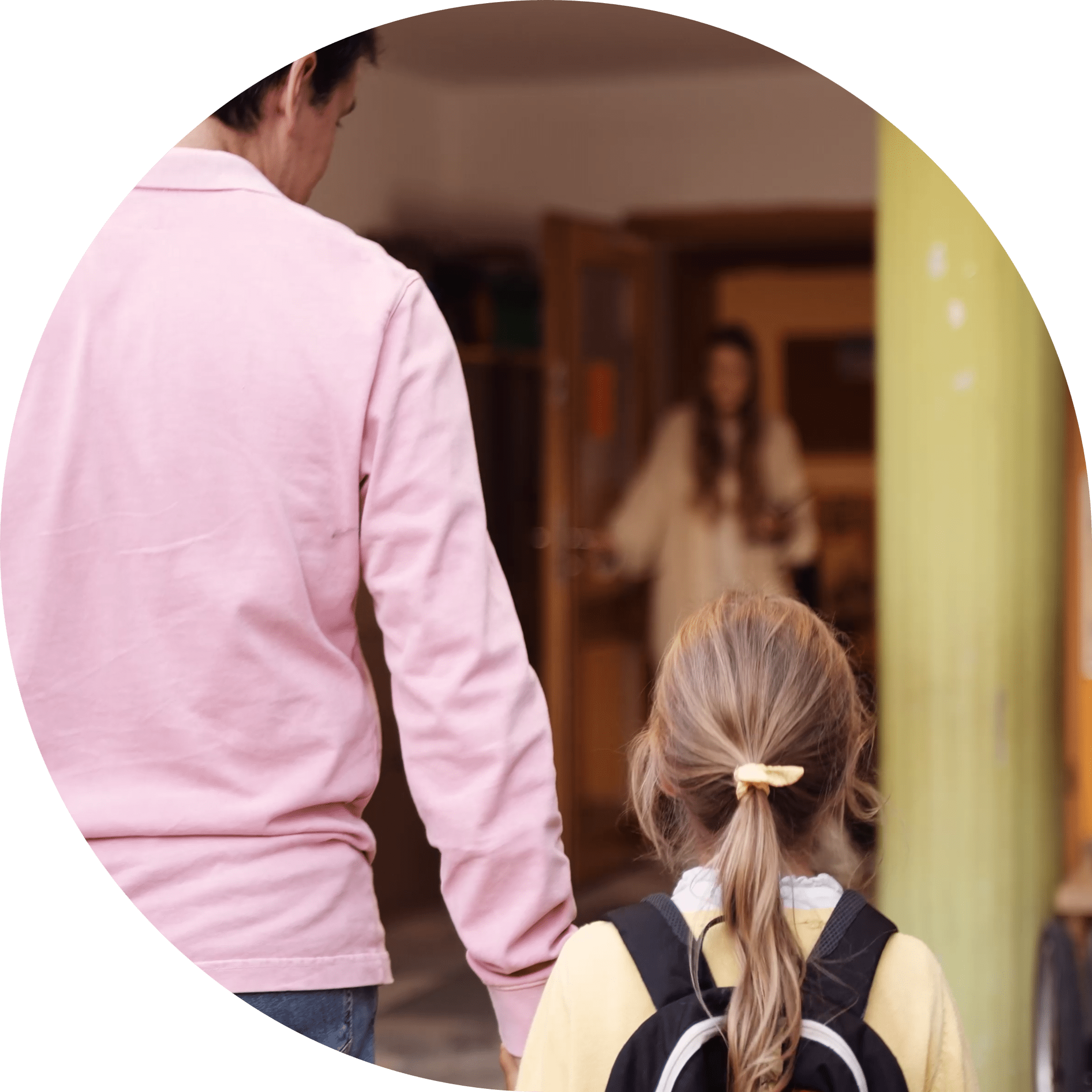 Vater hat seine Tochter an der Hand und ist auf dem Weg in den Kindergarten. Im Hintergrund sieht man die Erzieherin.